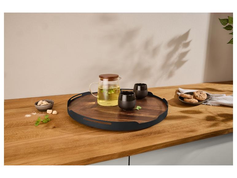 Houten dienblad met glazen theepot, zwarte kopjes, koekjes en suiker op een houten aanrecht.