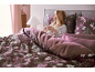 Vrouw in bed met bloemendekbedovertrek en kussens, drinkt thee.