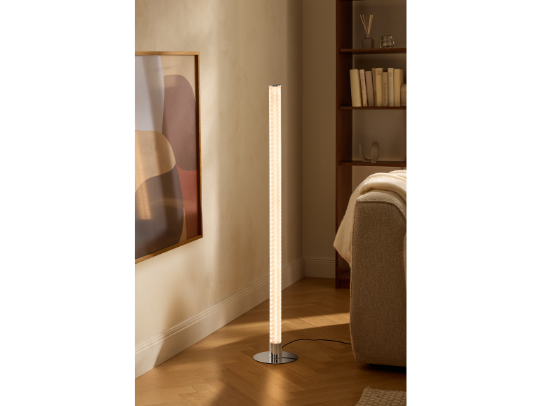 Cilindervormige vloerlamp met chromen voet verlicht een moderne woonkamer met boekenplank en bank.