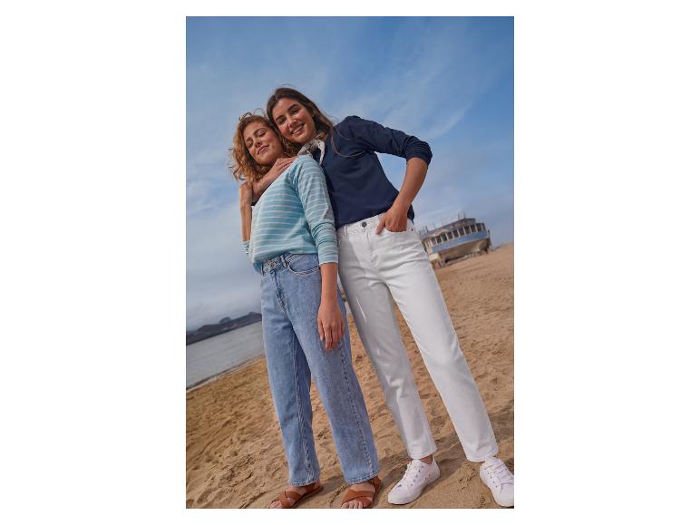 Twee vrouwen in jeans en shirts op het strand, met boot en zee op de achtergrond.