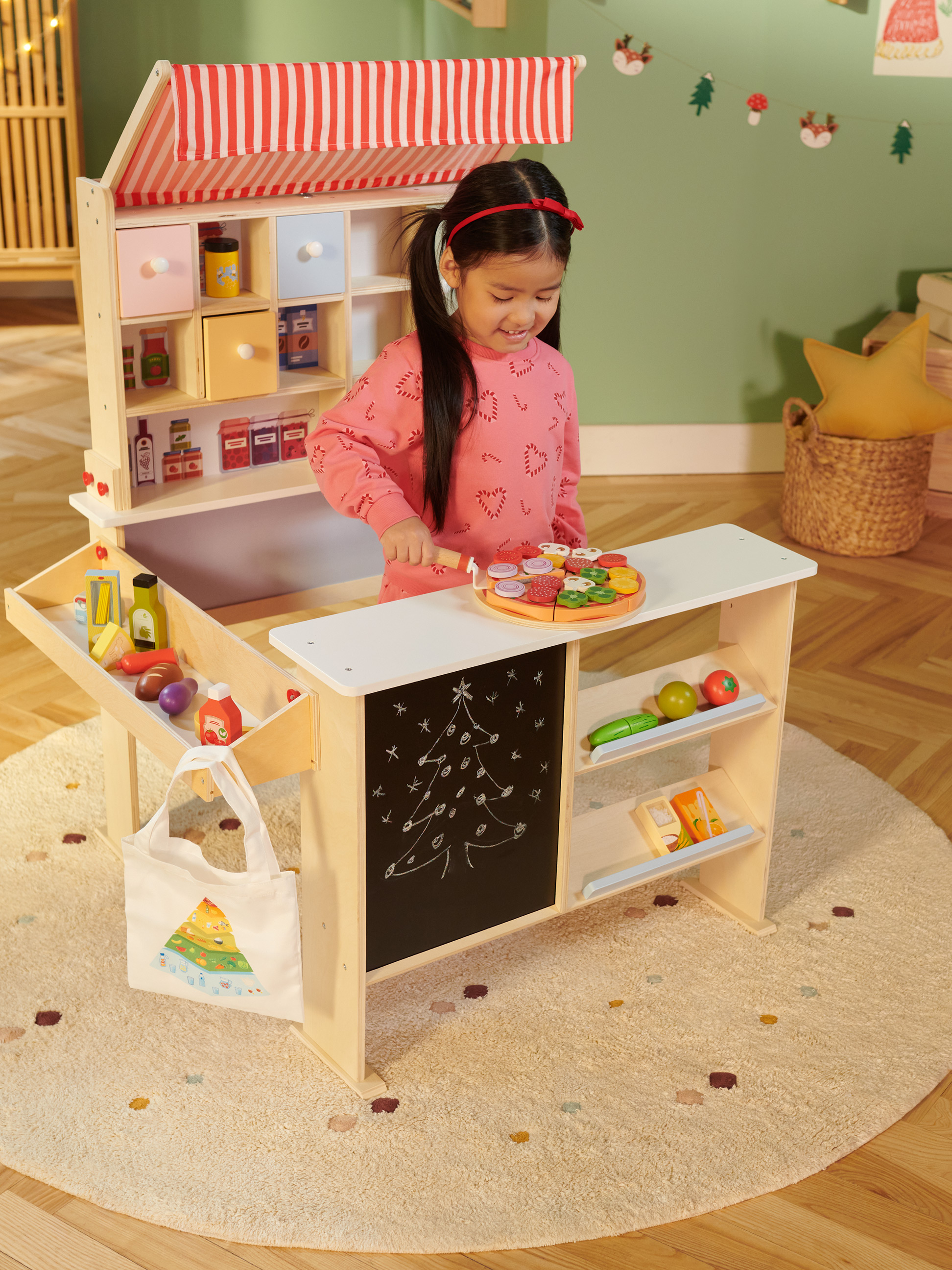 Lachend meisje speelt met een houten speelgoedwinkel, uitgerust met speelgoedeten en een krijtbord.