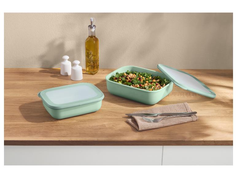 Twee mintgroene voedselcontainers, één met salade, op een houten aanrecht met olie en kruiden.