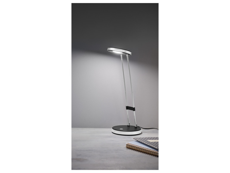 Een zilveren bureaulamp met een ronde lampenkap, op een bureau met een notitieboek.