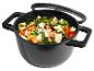 Zwarte pan met glazen deksel, gevuld met gesneden groenten zoals broccoli, wortels en paprika.