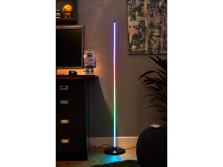 Een hoge, van kleur veranderende LED vloerlamp staat naast een donker bureau met een computermonitor.