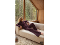 Vrouw in pruimkleurige loungewear ontspant op een bank bij een groot raam met uitzicht op een bos.