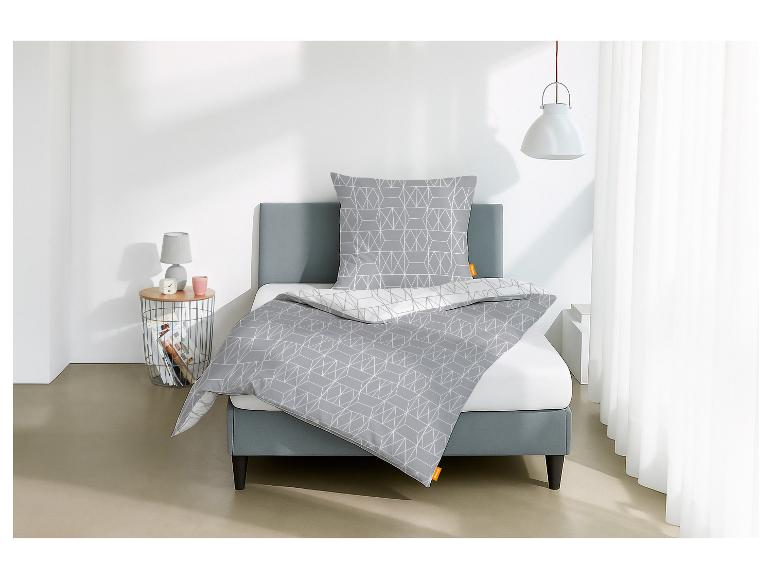 Grijs bed met geometrisch beddengoed, nachtkastje met lamp en witte gordijnen.
