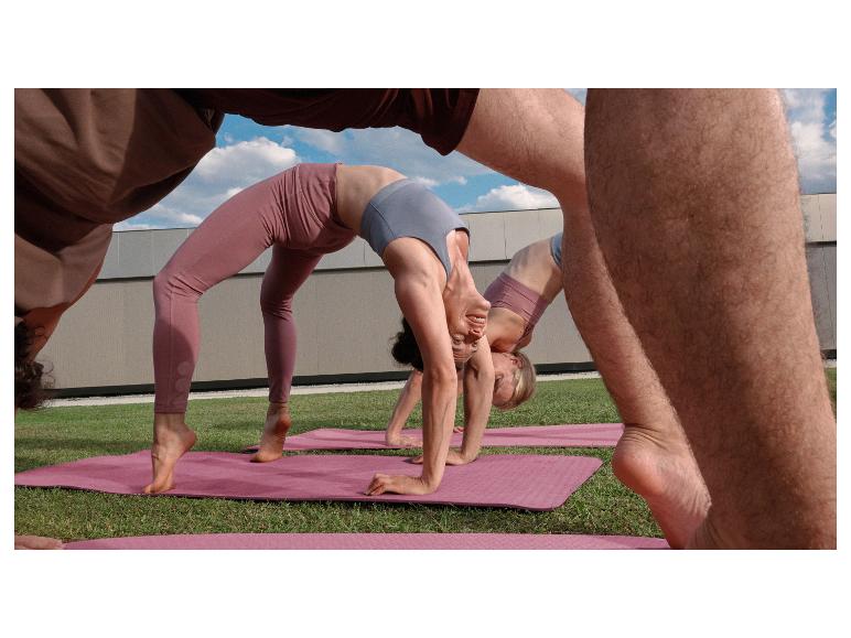 Drie mensen doen yoga op roze matten op gras, in leggings en sporttops.