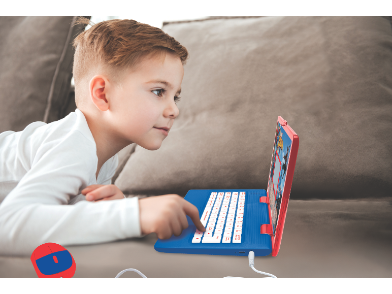Een jongen ligt op de bank en gebruikt een blauw-rode speelgoedlaptop.