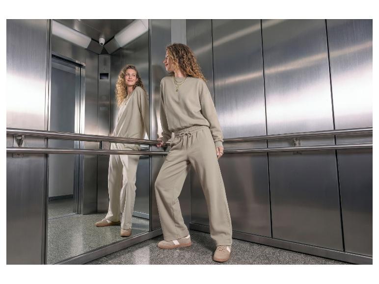Vrouw in een beige sweatshirt en joggingbroek, staand in een lift.