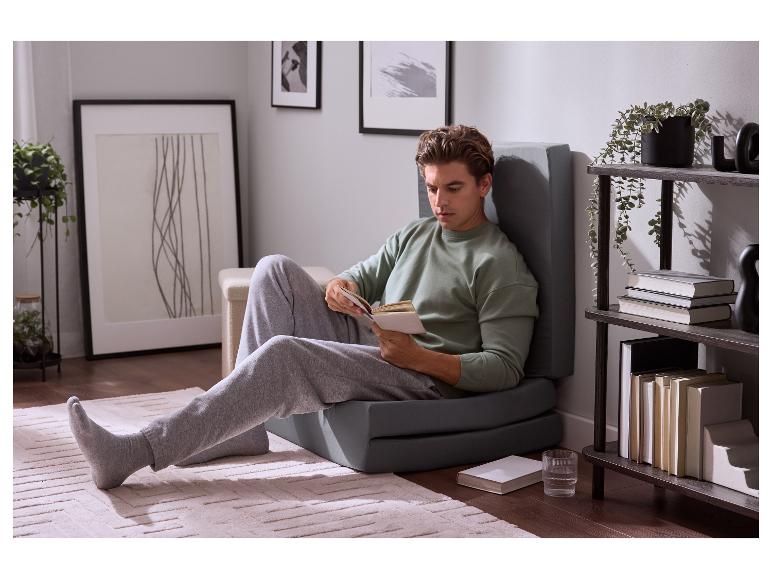 Man leest een boek, zittend op een grijs opvouwbaar matras in een woonkamer.