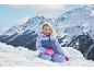 Lachend meisje in skipak en roze wanten in de sneeuw, met bergen op de achtergrond.