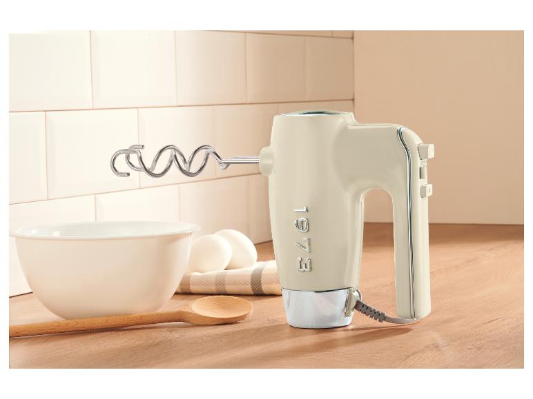 Crèmekleurige handmixer met deeghaken, kom, eieren en houten lepel op een aanrecht.