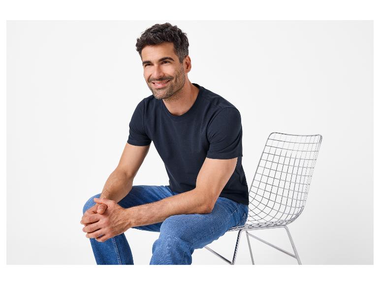 Man in donkerblauw T-shirt en jeans zit lachend op een stoel.