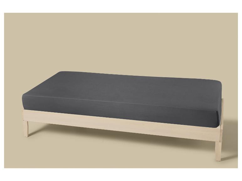 Grijs hoeslaken op een licht houten bed tegen een beige achtergrond.