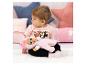 Baby in roze Minnie Mouse-top met een roze pop op een wit vloerkleed met kussens.