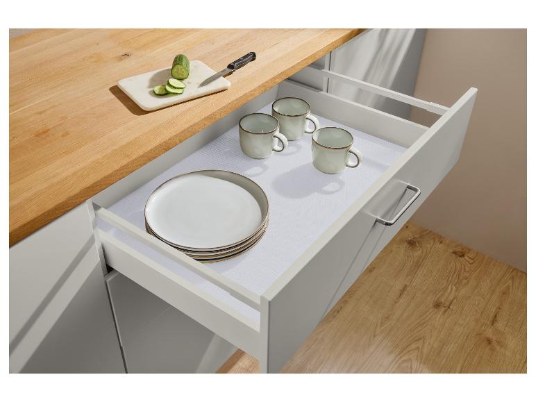 Open keukenlade met borden, mokken en antislipmat, met gesneden komkommer op het aanrecht.