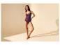 Vrouw in paarse lingerie, hemdje en slip, poseert tegen een beige achtergrond.