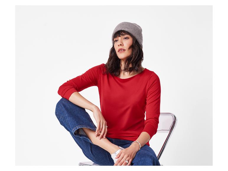 Vrouw in rode longsleeve, blauwe jeans en grijze muts zittend op een stoel.