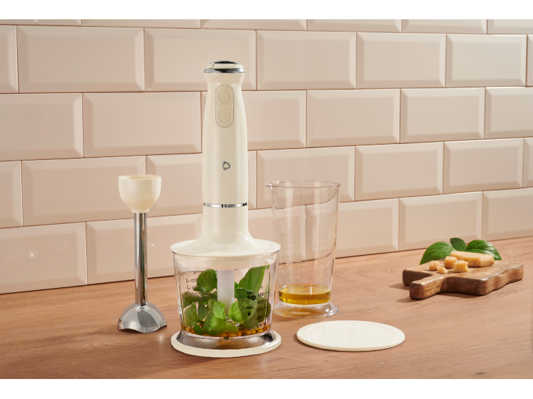 Crèmekleurige staafmixer met kruiden, olie en kaas op een houten oppervlak.