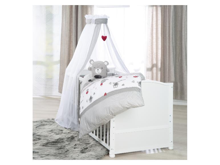 Witte babywieg met hemel, grijs geruit beddengoed en een pluche teddybeer.