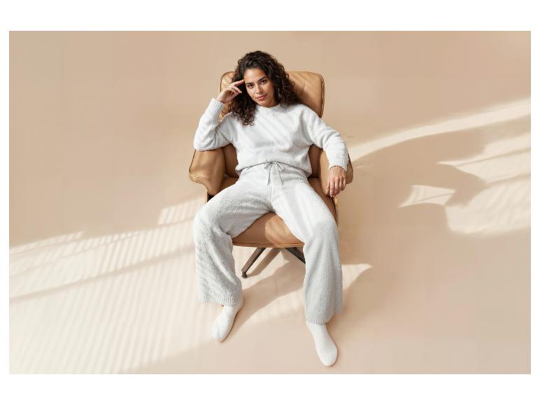Vrouw in een comfortabele witte loungewear set, zittend in een bruine fauteuil met raamlicht.