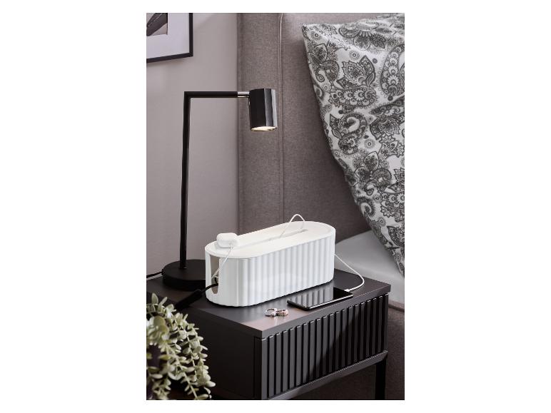 Zwart nachtkastje met een tafellamp, een witte kabelbox en een opladende smartphone.