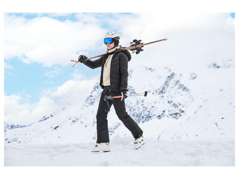 Vrouw in zwart-witte ski-uitrusting, met ski's op haar schouder, op een besneeuwde berg.