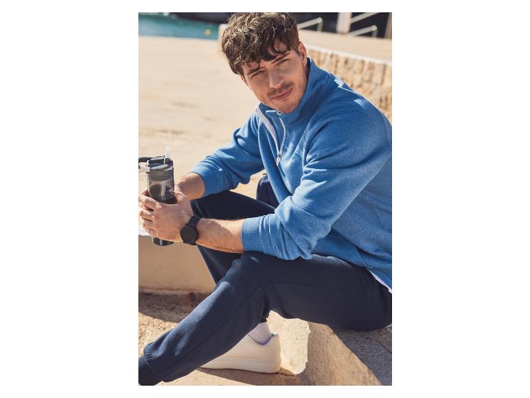 Een man draagt een blauwe trui en zwarte broek, terwijl hij een waterfles vasthoudt.