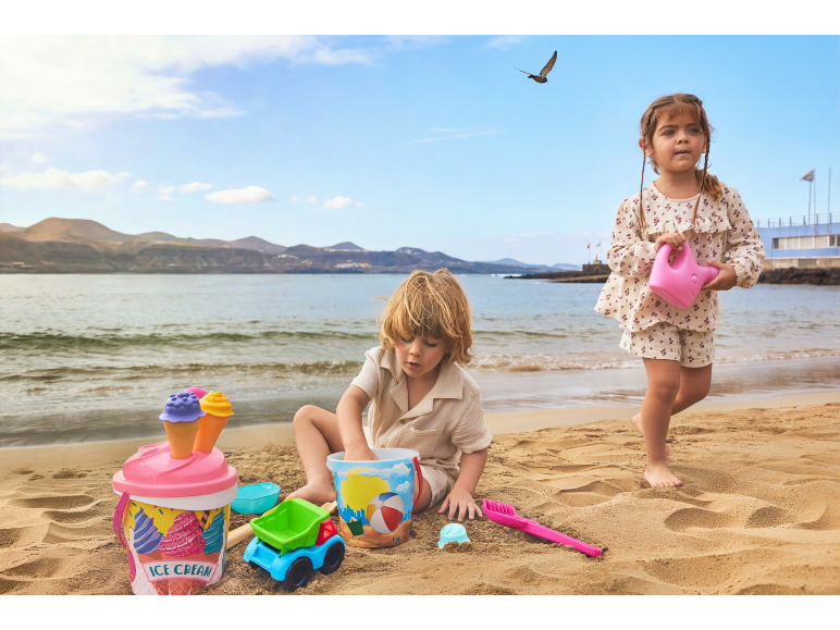 Twee kinderen spelen met zandspeelgoed, waaronder een ijsemmer en gieter, op het strand.
