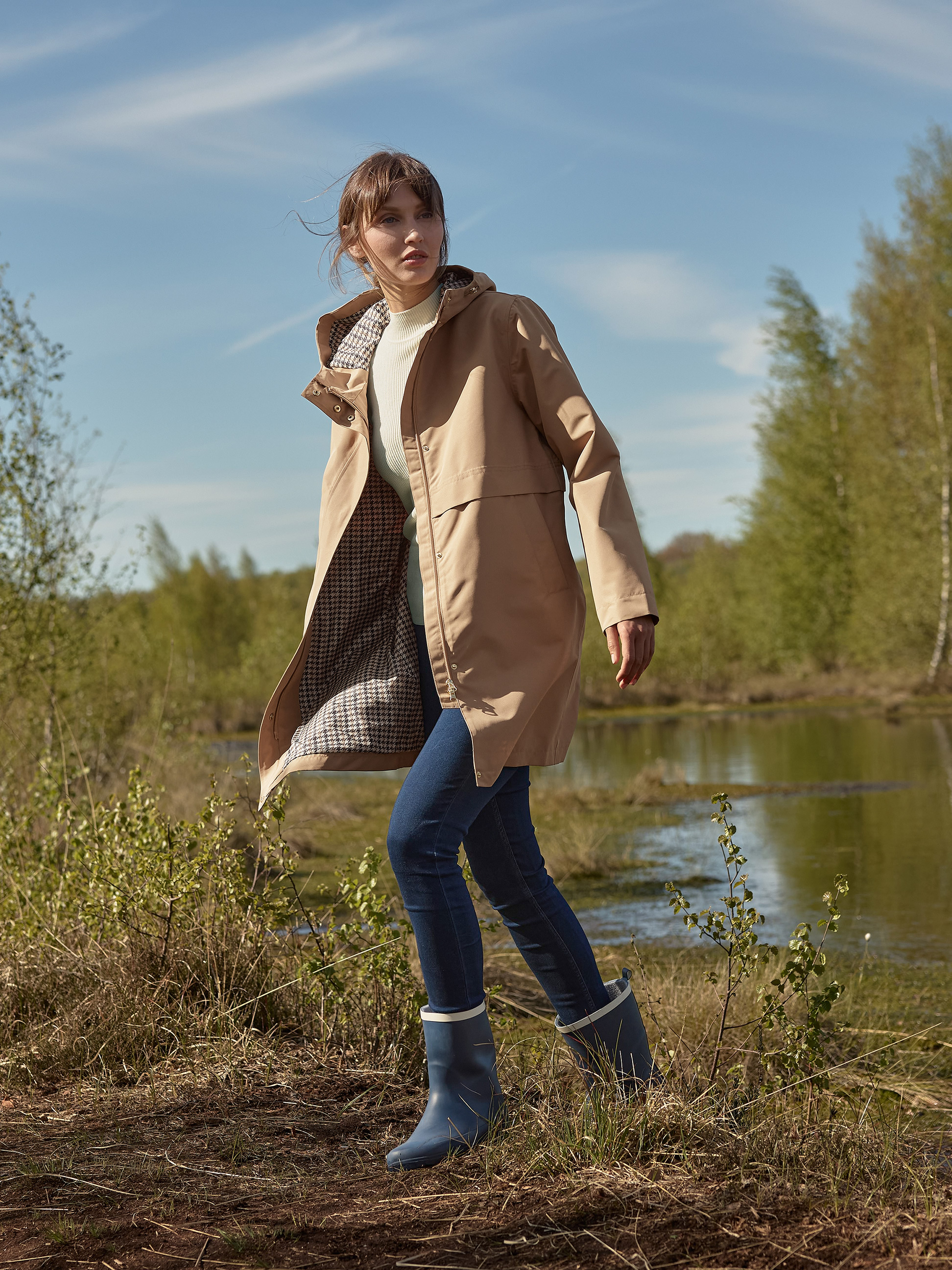 Vrouw in beige trenchcoat, jeans en blauwe regenlaarzen, lopend bij een meer.