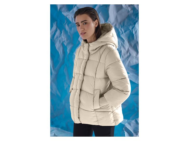 Vrouw in een beige gewatteerde jas met capuchon tegen een verfrommelde blauwe achtergrond.