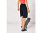 Een jongen draagt blauwe shorts, sneakers en een skateboard.
