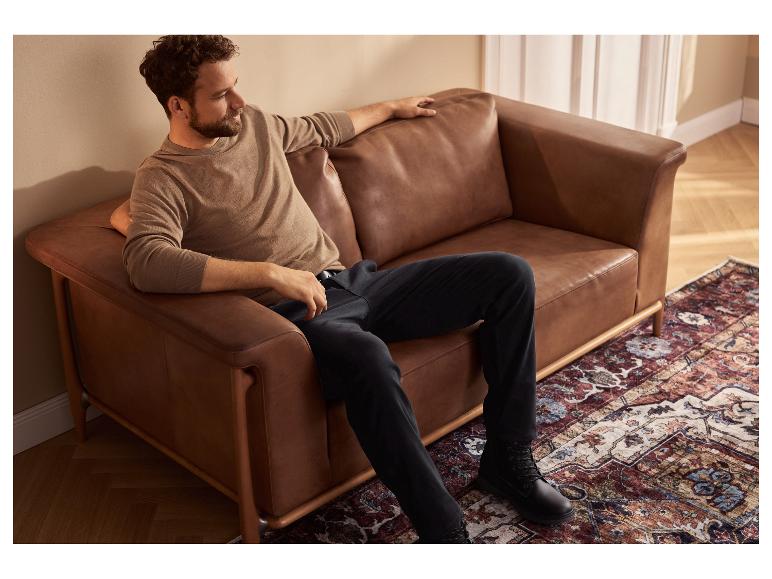 Een man zit op een bruine leren bank, gekleed in een beige trui en zwarte broek.