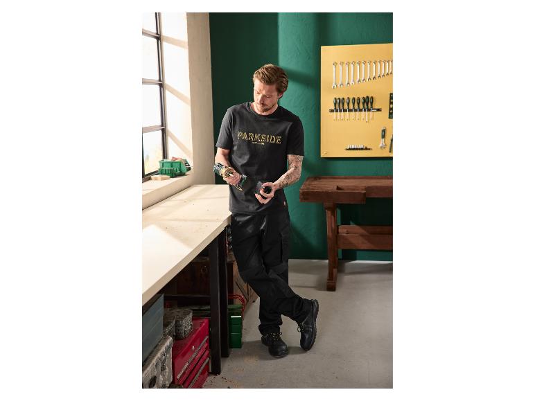 Man in zwart T-shirt en werkbroek, met een boormachine in een werkplaats.