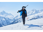 Man in ski-jas, broek, helm en bril draagt ski's en stokken op besneeuwde berg.