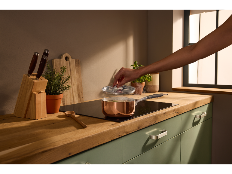 Keuken met houten aanrechtblad, groene kasten en een persoon die kookt met een koperen pan.