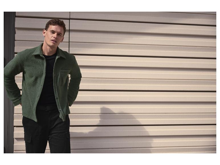 Man in een groen fleece overhemd en zwarte cargobroek, poserend voor een lamellenmuur.