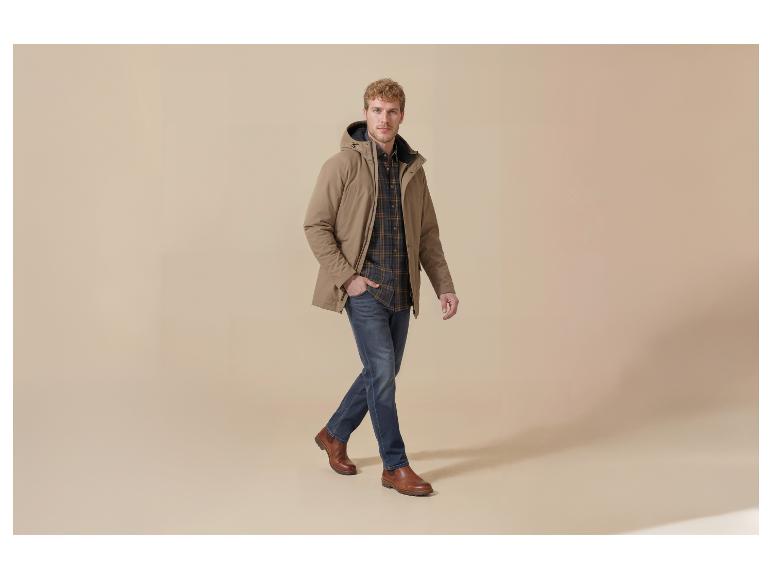 Man in beige jas, geruit overhemd, jeans en bruine schoenen, lopend.