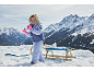 Meisje in ski-jas, broek en roze handschoenen, speelt met sneeuw naast een slee.