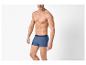 Man zonder shirt in blauwe boxershort, poserend tegen een witte achtergrond.