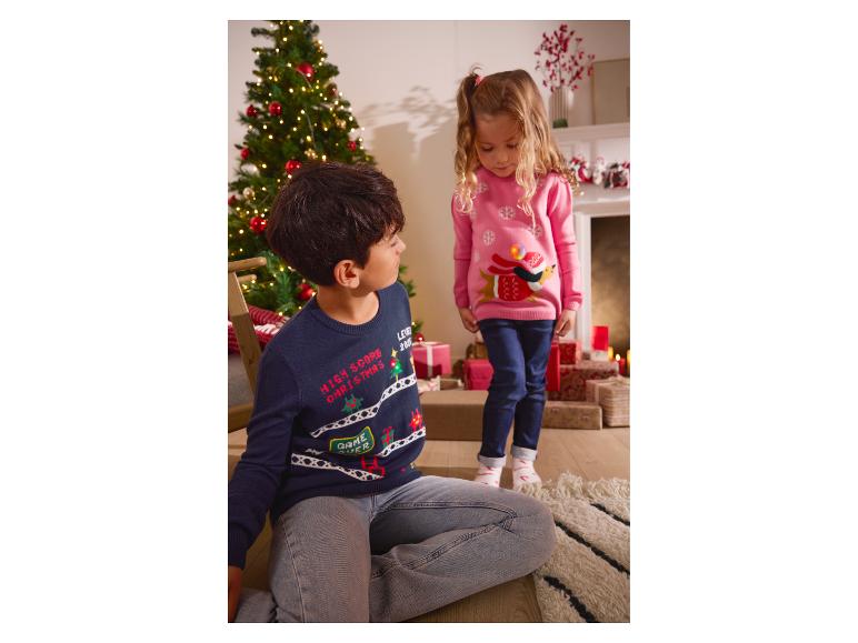 Twee kinderen in kersttruien, een jongen op de grond en een meisje staand, naast een kerstboom.