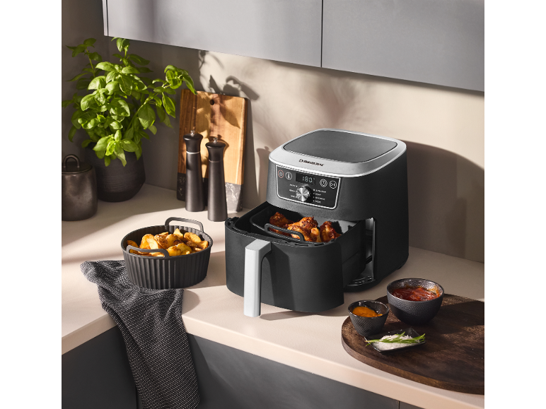 Zwarte airfryer op een aanrecht naast gebakken aardappelen en sauzen.