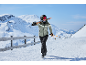 Vrouw in ski-jas, skibroek, helm en skibril draagt ski's en stokken in besneeuwde bergen.