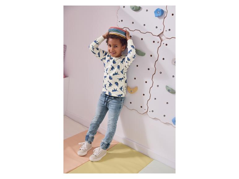 Een lachende jongen in een dinosaurus-top en jeans, staand bij een klimmuur.