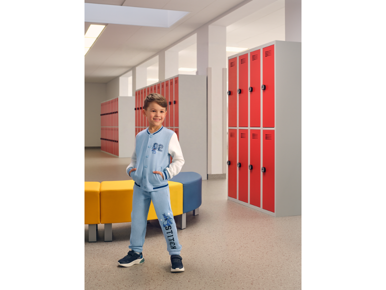 Jongen in lichtblauw trainingsjack en bijpassende broek in een schoolgang met rode kluisjes.
