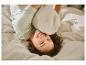 Lachende vrouw ligt op haar rug in bed met een beige dekbed en kussen.