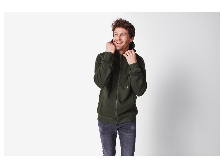 Lachende man in een donkergroene hoodie met rits en grijze jeans.