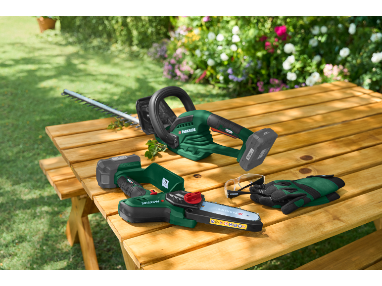 Accu heggenschaar en snoeizaag op een houten tafel in de tuin met veiligheidsbril en handschoenen.
