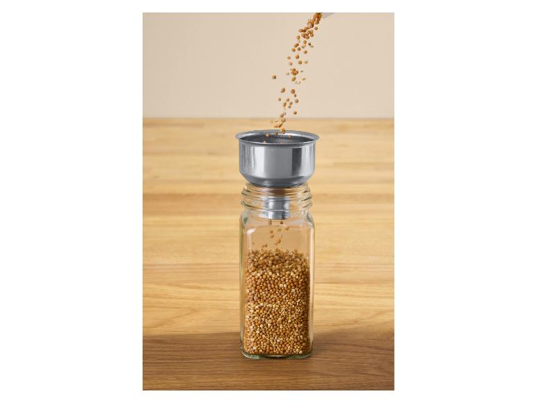 Gele mosterdzaadjes worden via een metalen trechter in een glazen pot gegoten.